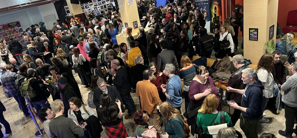 A large crowd gathers in a brightly lit indoor space, with many people chatting and holding drinks. A sign marked "CINE" is visible in the background.