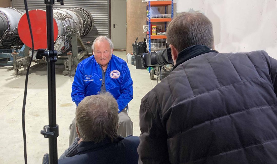A man in a blue jacket sits for an interview on camera in an industrial setting, with filming equipment and crew visible around him.