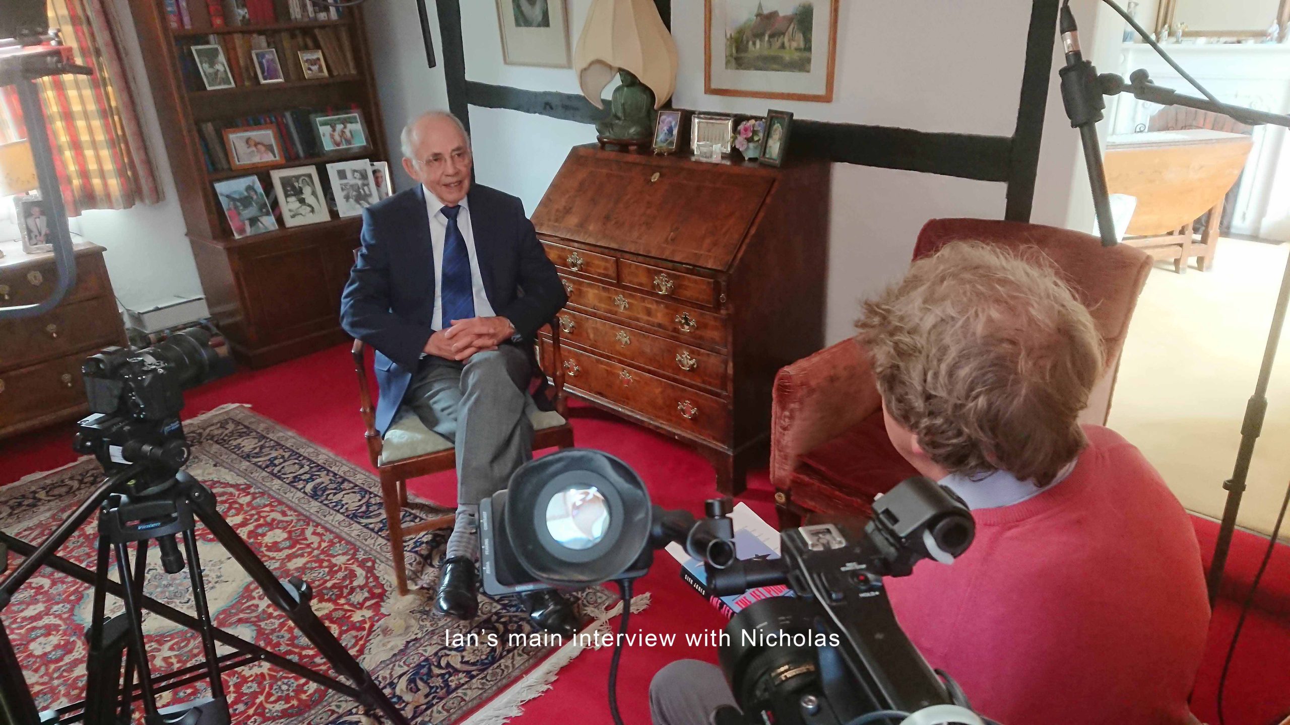 An older man in a suit sits for an interview in a cozy, carpeted room, facing another person with cameras set up around them.