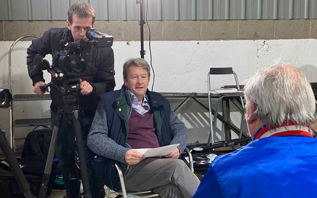 Three men are indoors; one is operating a camera, another is seated holding papers, and the third sits with his back to the camera. Filming equipment and chairs are visible.