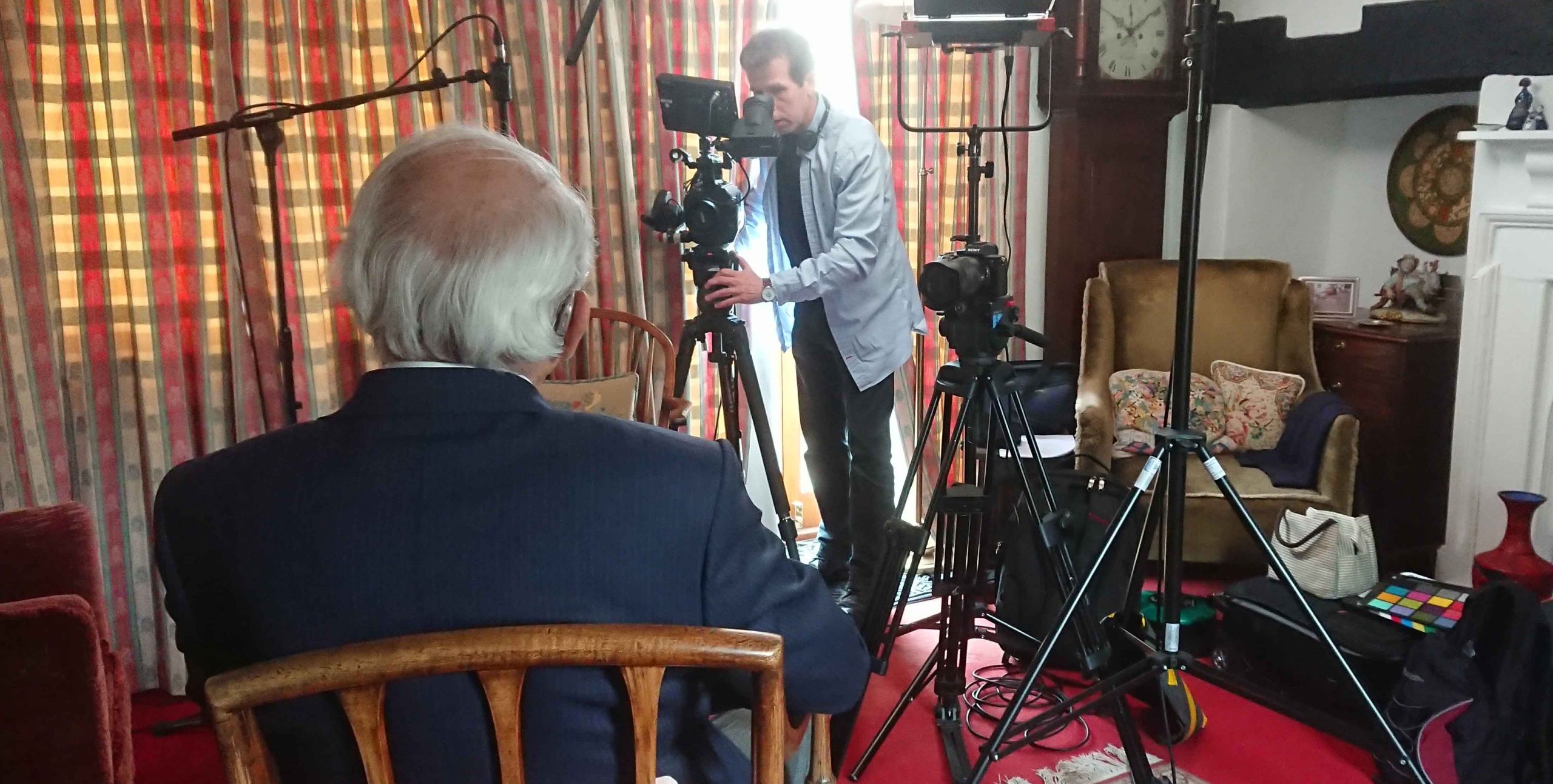 A man with gray hair sits facing camera equipment while another man adjusts a camera in a living room setup for an interview with lighting and microphones.